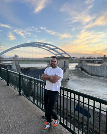 Shiv leaning on a railing above a river, bridge behind him